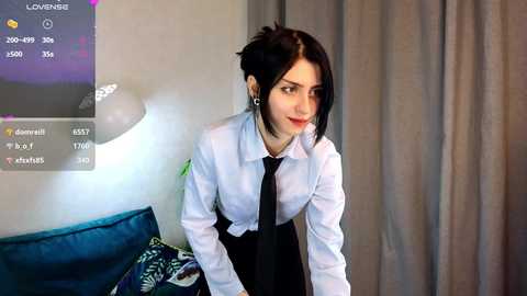 Media: Video of a young woman with pale skin and black hair, dressed in a white shirt and black tie, leaning forward in a dimly lit room.