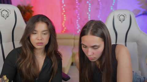 Media: Video of two young women with long brown hair, one in a black dress, the other in a sleeveless top, seated on gaming chairs. Background features colorful LED lights and a gaming setup.