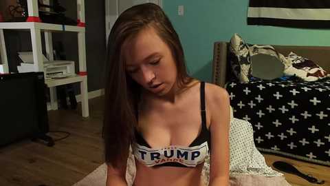 Media: Video of a young woman with long brown hair in a black \"TRUMP\" bra, sitting on a beige rug in a messy bedroom with a bed and teal walls.