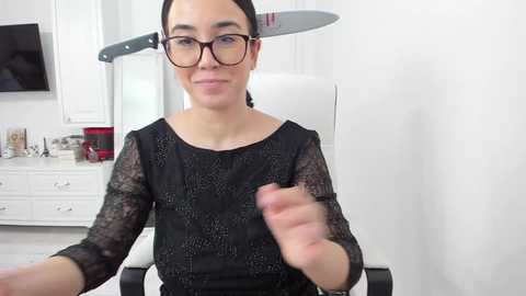 Media: Video of a light-skinned woman with dark hair, wearing glasses and a black lace top, holding a knife in a white room with white cabinets and a TV.