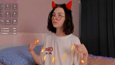 Media: Video of a young woman with shoulder-length dark hair, wearing red cat ears, round glasses, and a white t-shirt with a colorful graphic. She's holding glowing orange lights. Background shows a bed with blue sheets and a calendar with dates marked.