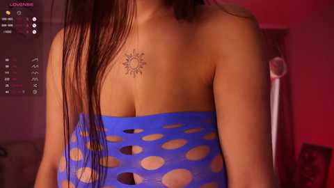 Media: Video of a woman with long brown hair wearing a blue, polka-dotted, strapless top, showcasing a sun tattoo on her collarbone. Background features a dimly lit room with red and pink hues.