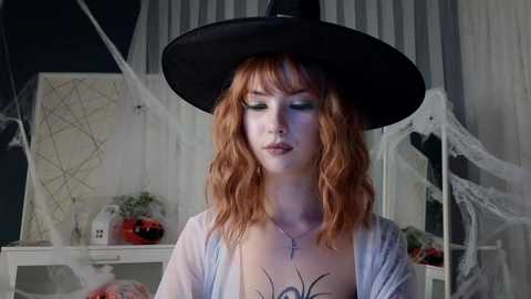 Media: Video of a pale, wavy-haired woman with green eyeshadow, wearing a black witch hat and sheer white robe, standing in a Halloween-themed room with spider webs and a pumpkin.