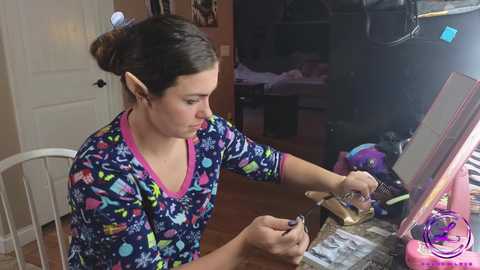 Media: A video of a young woman with long brown hair in a ponytail, wearing a colorful V-neck shirt, applying makeup at a cluttered vanity in a dimly lit bedroom.