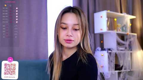 Media: Video of a young Asian woman with long, straight, light brown hair, wearing a black top, in a modern, cozy room with white shelves, and a teal couch in the background.