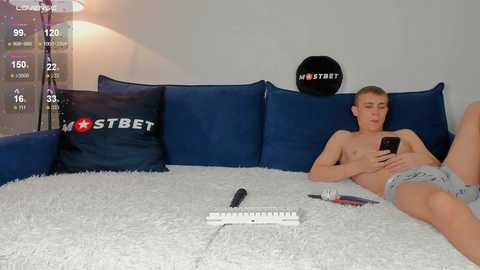 Media: Video of a young, shirtless, fit white man with short blonde hair lying on a plush white rug in a minimalist room with blue cushions and a \"HST\" pillow.