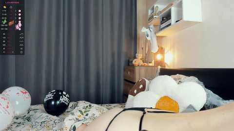 Media: Video of a cozy bedroom with a dark gray curtain, white shelves, a bedside table, and a stuffed animal on a bed with a floral-patterned sheet.