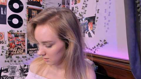 Media: Video of a young woman with long, blonde hair, wearing a white off-the-shoulder top, surrounded by a cluttered wall adorned with posters, records, and green vines.