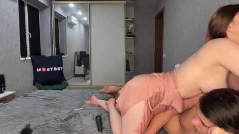 Media: Video of two Asian women, one topless in a pink skirt, performing oral sex on the other, in a dimly lit, grey-walled bedroom with a mirror, AC unit, and a green pillow on the floor.