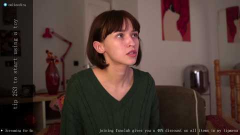 Media: Video of a young woman with short brown hair, wearing a dark green V-neck top, sitting indoors in a living room with wooden furniture, a red lamp, and abstract art on the wall.