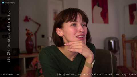 Media: Video of a woman with short brown hair, wearing a dark green top, sitting in a living room with a red vase and modern art on the wall.