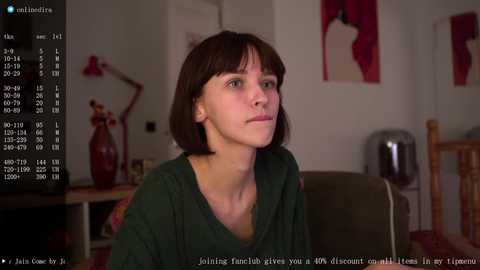 Media: Video of a pale, brunette woman with a short bob haircut, wearing a dark green V-neck sweater, seated indoors. Background includes a red lamp, a wooden chair, and a red vase. Text at bottom reads: \"Joining Tinder gives you a WMA with a 4.9 WNIS in the top 2%.\