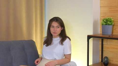 Media: Video of a young woman with long brown hair, wearing a white top, sitting on a gray couch in a modern living room with wooden walls and a potted plant on a black shelf.