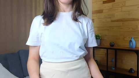 Media: Video of a woman with shoulder-length brown hair, wearing a white t-shirt and beige high-waisted pants, standing in a room with a wooden wall, blue sofa, and plant.