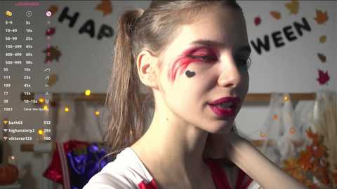 Media: Video of a young woman with fair skin and light brown hair styled in pigtails. She wears red lipstick and eye makeup, and a white shirt with red accents. Background shows Halloween decorations and \"HAPPY HALLOWEEN\" banner.