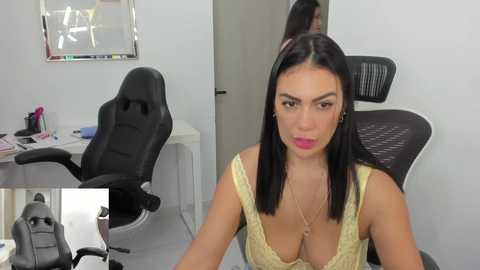 Media: Video of a woman with long black hair, wearing a low-cut yellow dress, sitting in a modern office with black ergonomic chairs and a white desk.