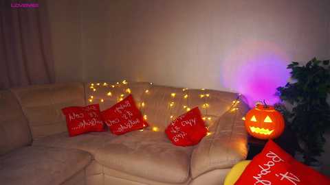 Media: A video of a cozy living room with a beige sectional sofa adorned with red pillows and warm yellow string lights. A lit jack-o'-lantern with a purple flame sits on the floor, adding Halloween charm.