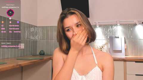 Media: Video of a young woman with shoulder-length brown hair, wearing a white lace camisole, hiding her face with her hands, in a kitchen with tiled walls and stainless steel appliances.