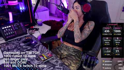 Media: Video of a tattooed woman with long dark hair, wearing camouflage shorts and a black tank top, using headphones, in a dimly lit room with Twitch chat visible.