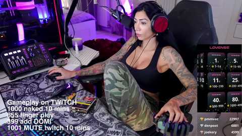 Media: Video of a tattooed woman in camo pants, black tank top, and headphones, sitting at a Twitch setup with a monitor displaying her stats, in a dimly lit room.