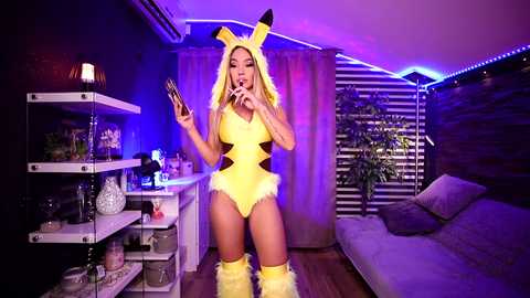 Media: Video of a blonde woman in a revealing Pikachu costume, holding a smartphone, standing in a modern living room with purple lighting, shelves, and a grey couch.