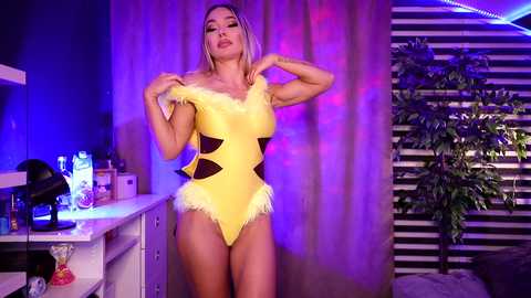 Media: Video of a blonde woman with a curvy physique, wearing a bright yellow, off-shoulder leotard with black geometric patterns, posing in a dimly lit room with purple and blue lights.