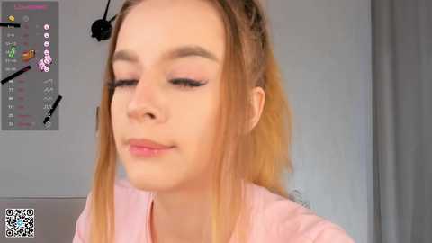 Media: Video of a young woman with light skin, blonde hair, wearing pink makeup, looking down, in a room with a gray wall and a calendar with colored markers.