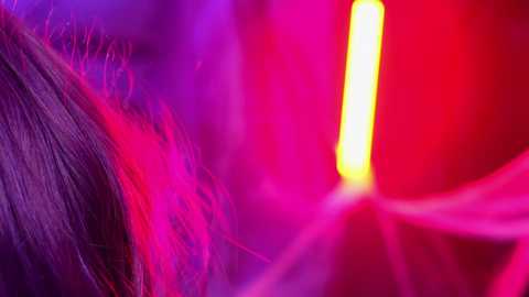 Media: Video of a close-up of a woman's hair, dyed in vibrant shades of purple, pink, and red. The background features a glowing, neon yellow light and blurred, intense red hues. The image evokes a modern, vibrant aesthetic.