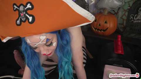 Media: Video of a woman with blue hair, dressed as a pirate, peeking under an orange pirate hat. She has a white patch over her eye and a skeleton face painted on her face. Background features a jack-o'-lantern and a red candle.