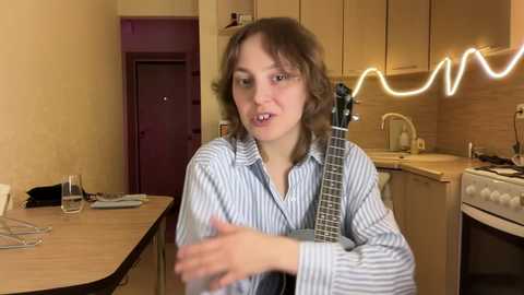 Media: Video of a young woman with shoulder-length brown hair, wearing a striped shirt, playing a guitar in a cozy kitchen with a neon sign.