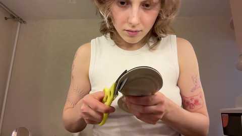 Media: Video of a young woman with light brown, wavy hair wearing a white tank top. She uses yellow-handled scissors to cut open a metal can. Background features a plain beige wall and a partially visible window with a curtain.