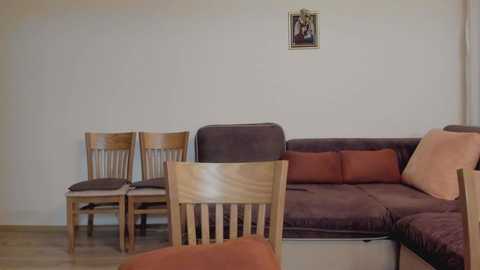 Media: A video of a minimalist living room with beige walls, featuring a dark brown L-shaped couch, wooden chairs, and a small framed picture above the couch. The floor is light-colored wood.