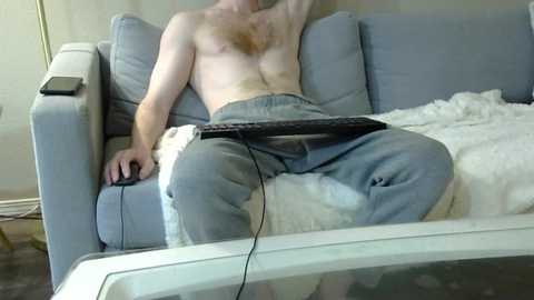 Media: A topless, light-skinned man with a hairy chest lounges on a light blue couch, wearing loose grey sweatpants, with a black remote control in hand. A white blanket and a smartphone are nearby.