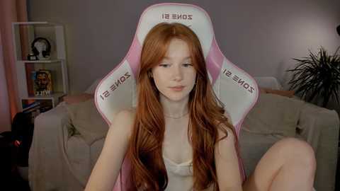 Media: Video of a young red-haired woman in a white lace bra, seated in a pink gaming chair in a cozy, dimly lit living room with beige furniture and a bookshelf.