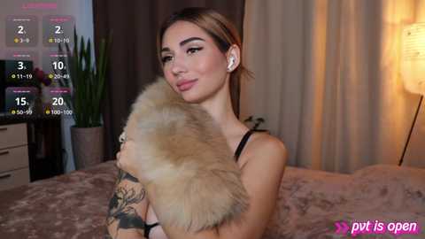 Media: Video of a light-skinned, tattooed woman with brown hair and a plush beige fur jacket, sitting on a bed with a beige blanket, in a dimly lit room.