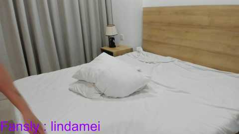 Media: A video of a modern, minimalist bedroom featuring a neatly made bed with white linens, a wooden headboard, beige curtains, and a small bedside table with a lamp.