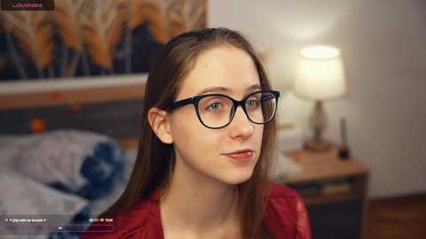 Media: Video of a fair-skinned woman with long brown hair, wearing black-framed glasses and a red top, sitting indoors, with a blurred background featuring a bed, lamp, and abstract wall art.