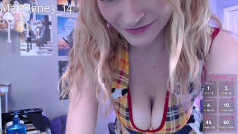 Media: A video of a blonde woman with wavy hair, wearing a low-cut, yellow plaid top, smiling, in a cluttered room with posters and a video game controller.