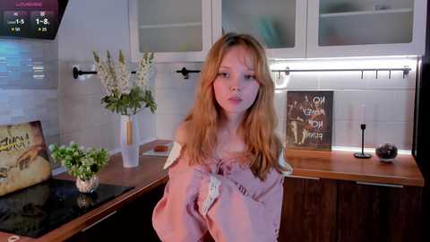 Media: Video of a young blonde woman in a pink off-shoulder dress, standing in a modern kitchen with white cabinets, wooden countertops, and floral decorations.