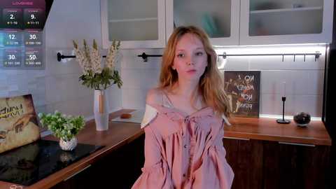 Media: Video of a young woman with long blonde hair, wearing a pink off-shoulder dress, standing in a modern kitchen with white tiles, wooden counter, and decorative items.