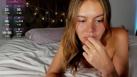 Media: Video of a topless woman with long brown hair, lying on a bed, biting her finger, in a dimly-lit room with a tufted headboard and string lights.