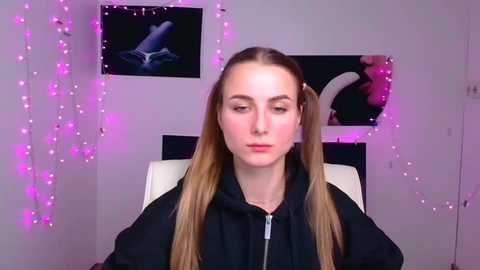 Media: A video of a young woman with long, straight blonde hair styled in pigtails, wearing a black hoodie, seated indoors. The background features a pink-lit wall with a black and white framed photo of a nude woman and fairy lights.