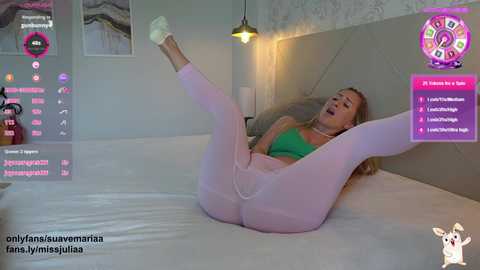 Media: Video of a blonde woman in green bra and light pink leggings, lying on a bed, legs raised, eyes closed, surrounded by virtual reality gaming interface.