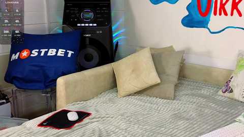 Media: Video of a cluttered bedroom with a beige bed, a Sony speaker, a blue \"MOSTBET\" pillow, a white mouse, and a blue wall with \"OIRK\" graffiti.