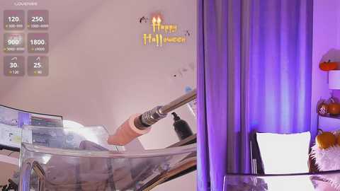 Media: Video of a modern bedroom with purple curtains, a cozy bed with white pillows, and a \"Happy Halloween\" sign. A person is using a steamer on a glass table.