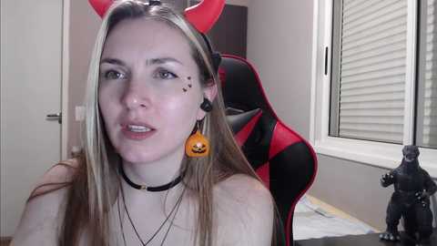 Media: A video of a young woman with fair skin, wearing a red devil horns headband, black choker, and an orange Jack-o'-lantern earring. She sits in a red and black gaming chair in a room with a window and a toy figurine.