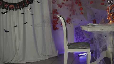 Media: Video of a Halloween-themed room with white curtains, bat banner, white chair, and round table adorned with spider decorations, purple lighting, and festive red and orange background.