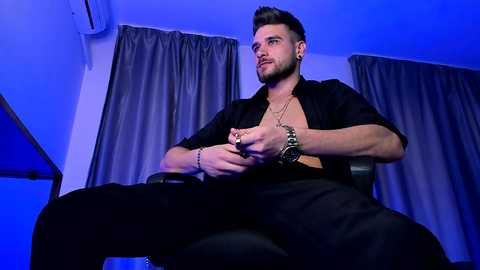 Media: Video of a young, muscular, bearded man with styled hair, wearing a black shirt, sitting with legs spread, against a blue-lit background with grey curtains.