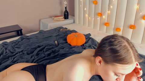 Media: A video of a fair-skinned woman with brown hair tied back, lying topless on a bed, wearing black underwear, with a pumpkin and string lights in the background.