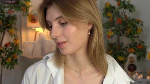 Media: A video of a blonde woman with shoulder-length hair, wearing a white shirt and gold necklace, standing in a brightly decorated room with floral arrangements.